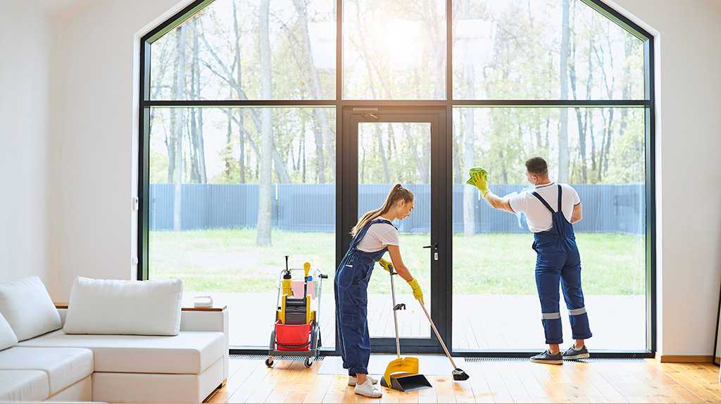 Two cleaners wipe down the window and sweep the floor with a broom. Moving Help makes home cleaning easy when you can hire the same moving labor company to clean your home and unload your belongings.