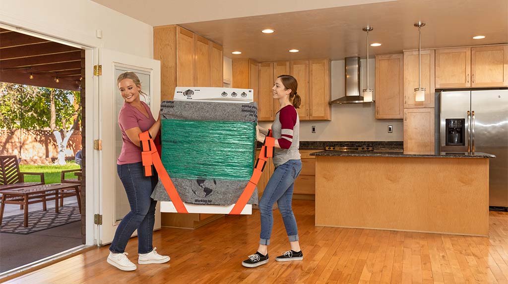 Two women carry a washing machine using moving equipment to easily load it to a truck using orange lifting straps.
