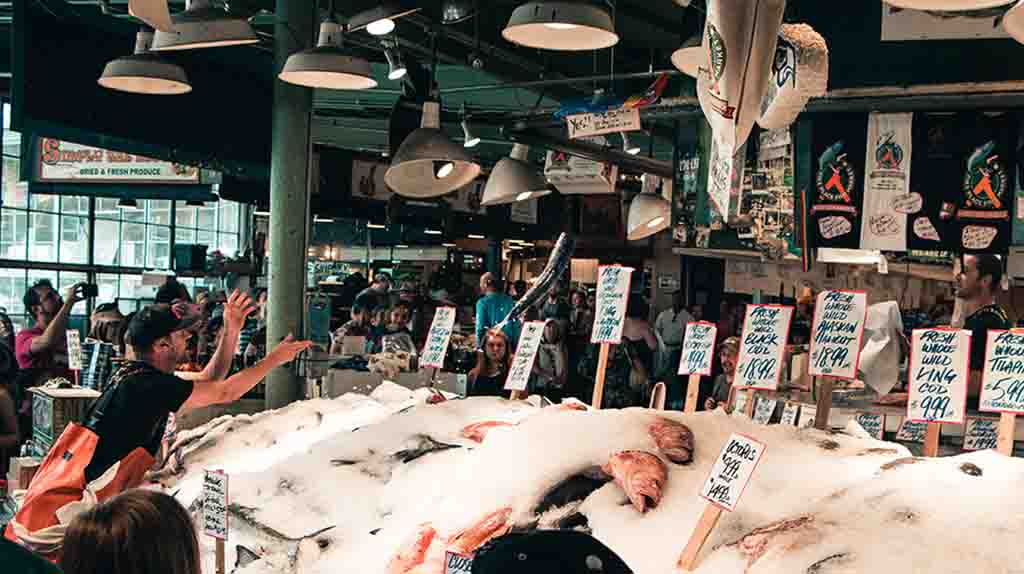 A man tosses a fish to another man at the Pike Place Market. When visitors go to Pike Place Market, they can watch the famous in-person fish throwing scene.