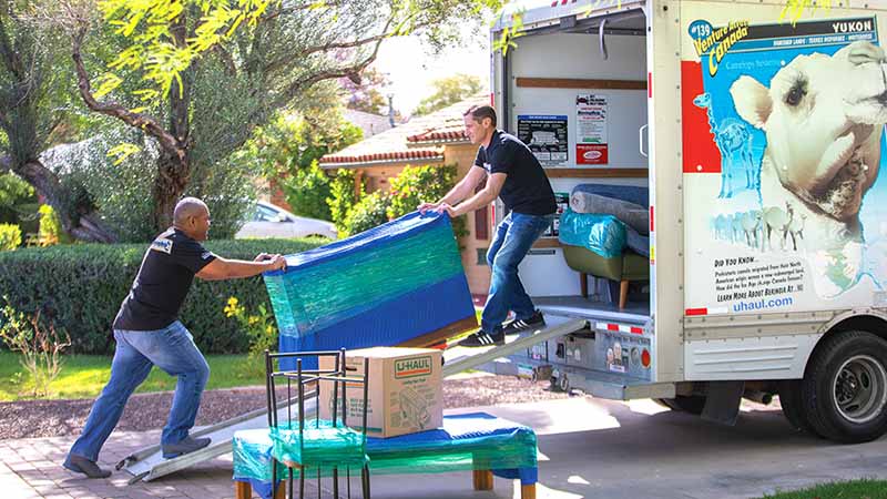 A moving labor team pushes a wrapped piece of furniture up a moving truck ramp.