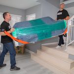 Two local moving labor providers carry a wrapped couch up a set of stairs.