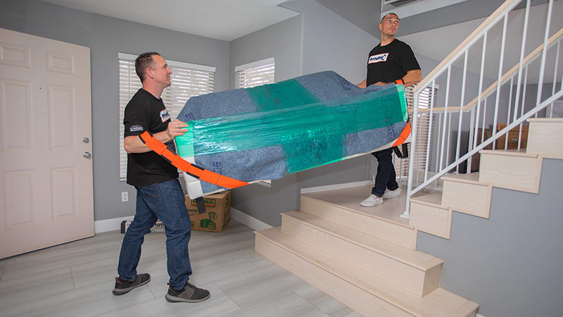 Two local moving labor providers carry a wrapped couch up a set of stairs.