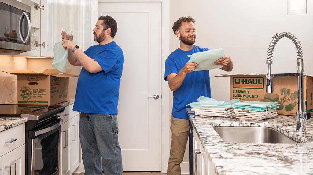 Two Moving Help Service Providers pack dishes into cushions and then into kitchen moving boxes for a customer.