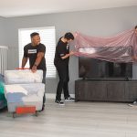 Two Moving Help Service Providers prepare to move a television by covering it with protective wrap. Another provider moves living room boxes with a dolly.