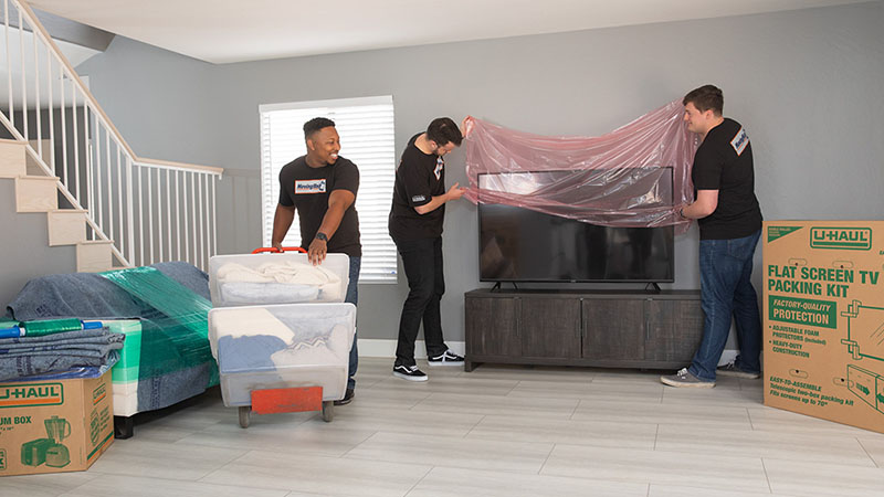 Two Moving Help Service Providers prepare to move a television by covering it with protective wrap. Another provider moves living room boxes with a dolly.