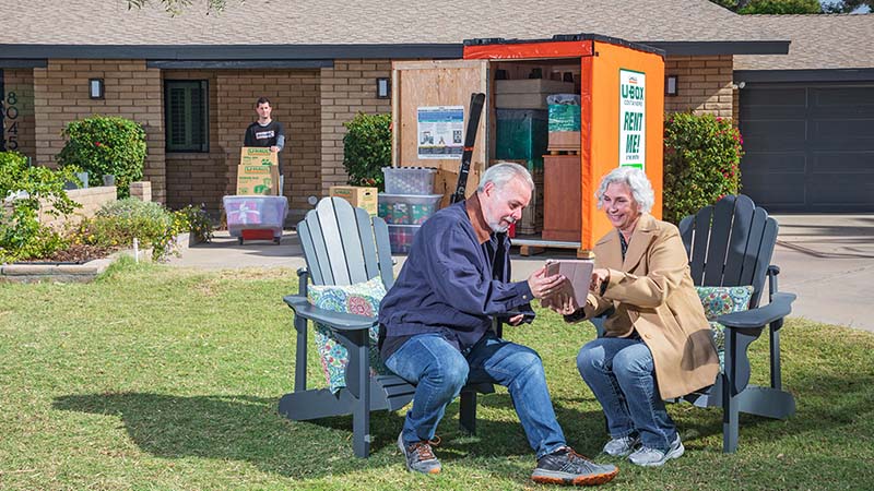 An older couple sits by a U-Box storage container as a Moving Helper in the background brings moving boxes on a dolly into the container.