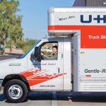A woman starts driving a U-Haul truck rental out of the parking lot.