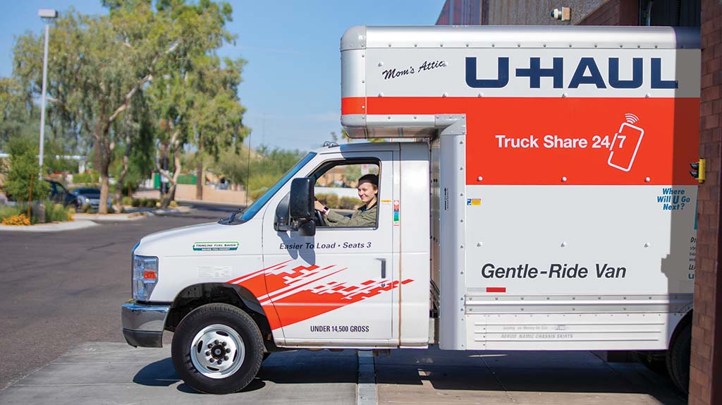 A woman starts driving a U-Haul truck rental out of the parking lot.