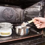 A person holds a paint brush to clean the inside of their oven using a mixture of baking soda and water that creates a paste. A jar of baking soda and a mixing bowl are sitting inside the oven.