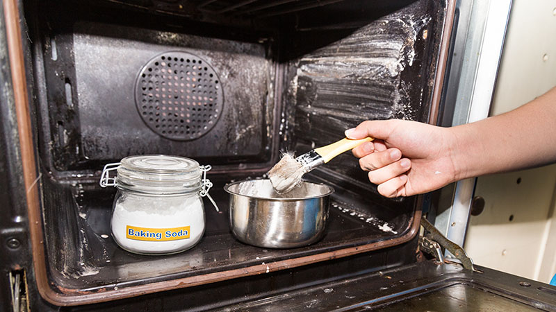 A person holds a paint brush to clean the inside of their oven using a mixture of baking soda and water that creates a paste. A jar of baking soda and a mixing bowl are sitting inside the oven.