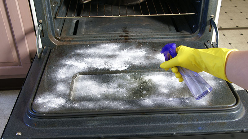 A person sprays white vinegar onto a baking soda paste to let the solution foam on the oven.