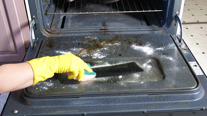 A person wipes the inside of the oven’s glass window using a sponge to clean the rest of the baking soda paste solution.