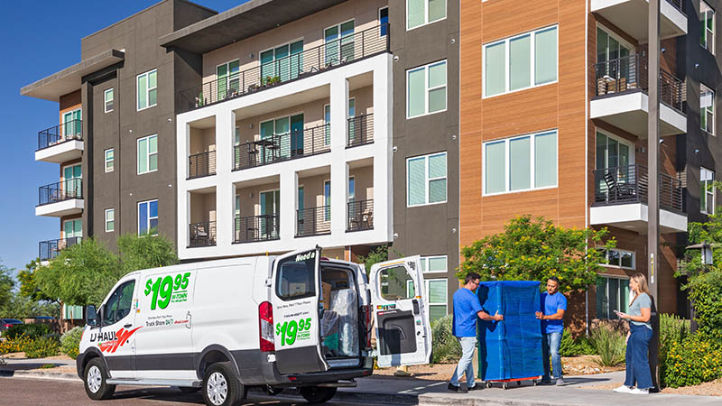 Two Moving Help Service Providers unload a tall cabinet using a dolly out of a U-Haul van rental for a female customer outside of a multifamily apartment complex.