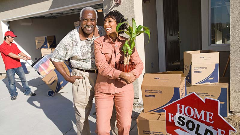 A couple stands in front of a house they recently bought together. In the background, a mover pushes a dolly with moving boxes into their home.