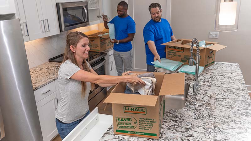 Two Moving Helpers pack kitchen items into moving boxes while a customer packs her dish towels into a moving box.