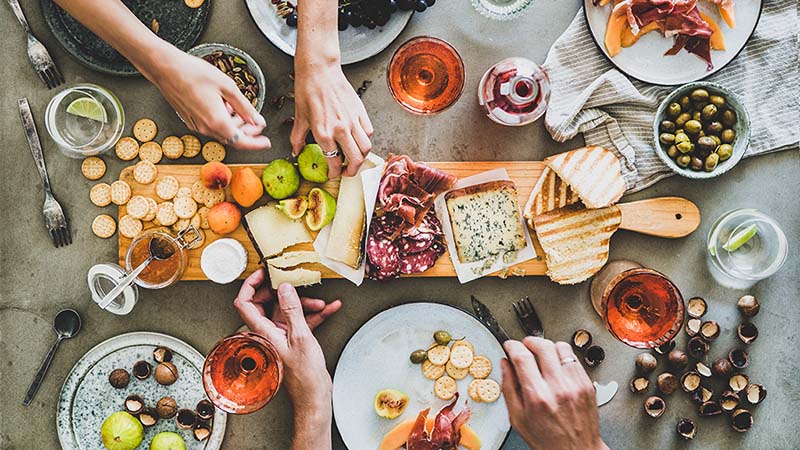 A group of friends sit around a charcuterie board as they begin piling ingredients from the board onto their plates. A charcuterie board experience could be a great going away party idea.