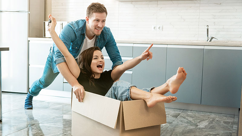 A man pushes a woman sitting in a moving box as they celebrate being in their new home.