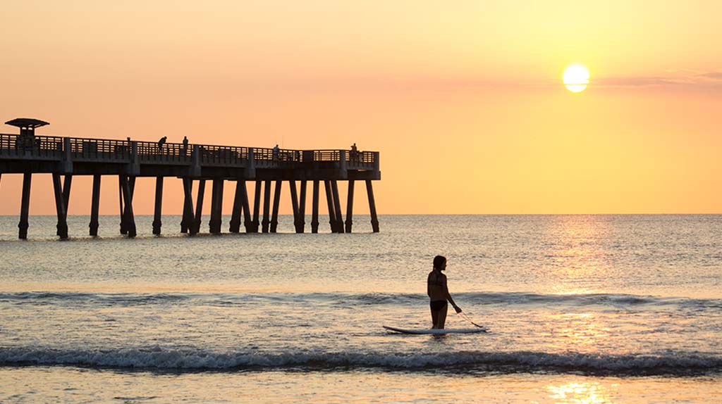 A woman stands in the water next to her paddleboard on one of Jacksonville’s beaches as the sun begins to set. Jacksonville has more shoreline than any other city in the nation along with 1,100 miles of navigable water.