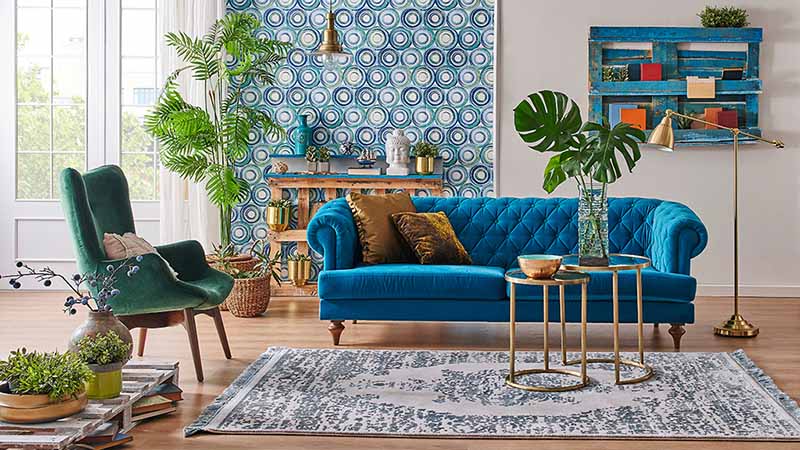 A blue couch sits in the living room with a blue rug, a green armchair, plants, other decor, and blue wallpaper in the background. DIY and Decor blogs are a great way to find new ideas on how to redecorate certain rooms in your home.