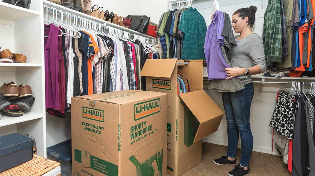 A woman takes her clothes with hangers and places them inside a U-Haul wardrobe moving box that has a metal rack installed already in the box. This way, she can quickly load and unload her clothes with hangers.
