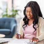 A woman sitting in her chair is going over her tax information as she holds a pen near a piece of paper while she uses a calculator and a computer to help her.