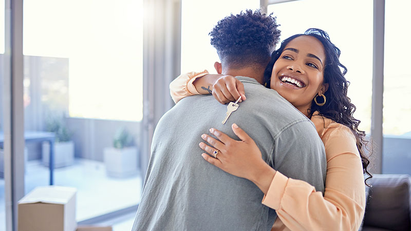 A woman hugs a man as she’s holding the key to their new apartment. Most landlords don’t care whether your lease start date vs. move-in date is on the same or different day.