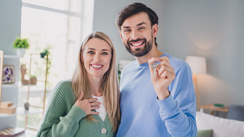 A woman and a man stand together smiling as the man holds their apartment’s key as they stand inside their new apartment.