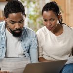 A man and a woman sit together on a couch as they review a printed bill and their finances on a computer. The cost of living varies between states and cities.