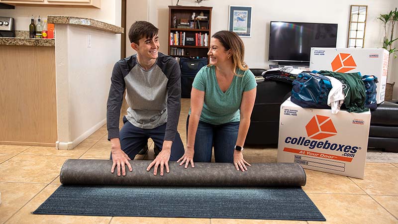 A mother helps a son roll a rug from their house for the son to take with him for college.