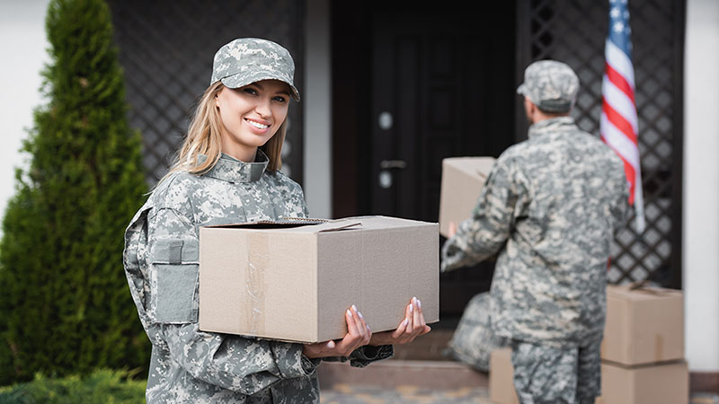 A woman in the military holds a moving box while her husband, who is also in the military, carries a moving box into their new home.