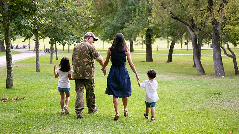 A man in the military holds his wife’s hand with one hand and his daughter’s hand with the other. The woman holds their son’s hand with her free hand as they all walk together in a park. The answer to “how often do military families move” is about 2.5 years.