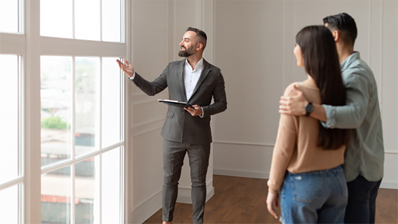 A realtor shows a couple the large window with an outside view in a living room of a potential house they may purchase.