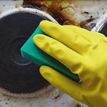A person wears cleaning gloves while using a sponge to clean a dirty electric stovetop.