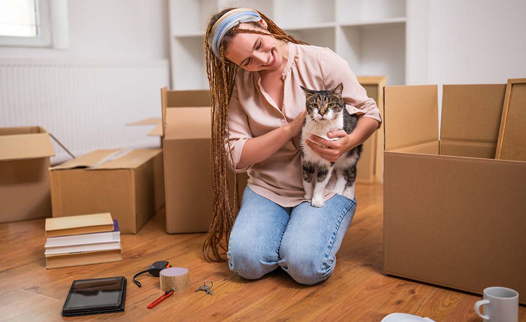 A woman holds her cat in her new home with moving boxes surrounding them on the hardwood floor. Moving with pets takes careful planning.
