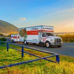A U-Haul truck rental drives down an empty, fall road while towing a U-Haul cargo trailer.