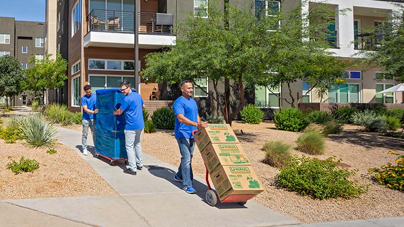 A labor-only mover pushes a utility dolly while carrying three small U-Haul moving boxes. Meanwhile, two local, labor-only movers, who are behind the first helper, push a piece of furniture on a furniture dolly.