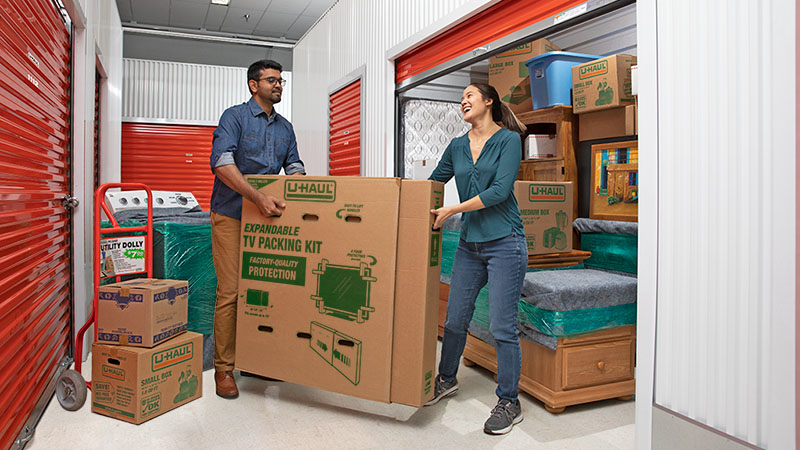 A man and a woman carefully move a TV moving box into their climate-controlled storage unit.