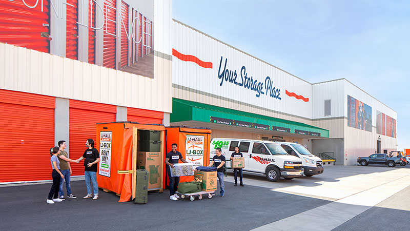 Three Moving Help Service Providers carefully load items into a U-Box storage container. The fourth moving labor Service Provider shakes hands with a man as he talks to a man and a woman.