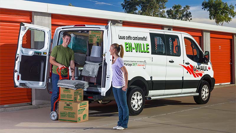 A man and woman smile at each other as they begin unloading their belongings out of a U-Haul cargo van and into an outdoor/drive-up storage unit.
