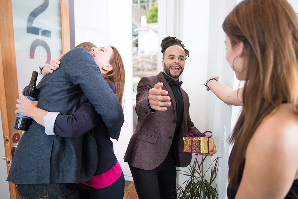 A couple hugs another couple at their front door as the couple invited to the housewarming party brought a gift for the couple’s party.