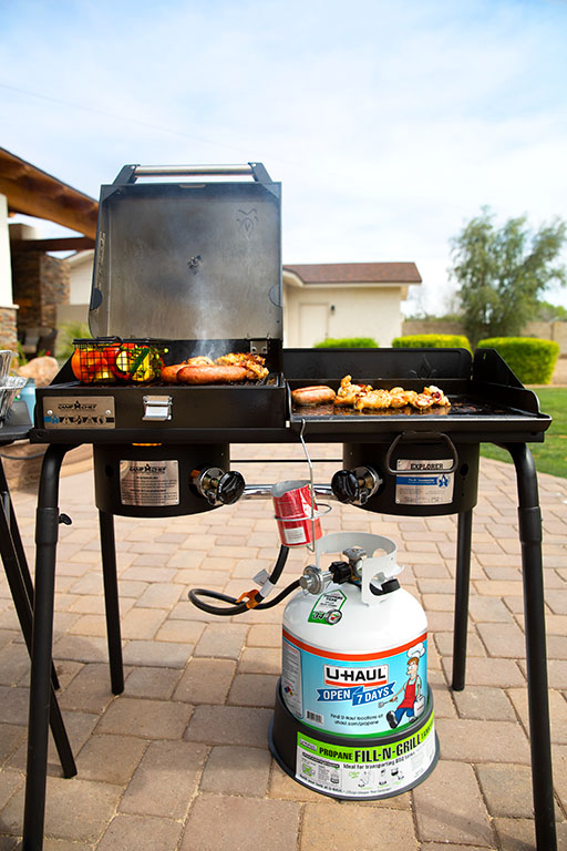 An outdoor grill is cooking sausage, corn, kebabs, and vegetables while using a U-Haul propane tank at a housewarming party for a new family’s home.