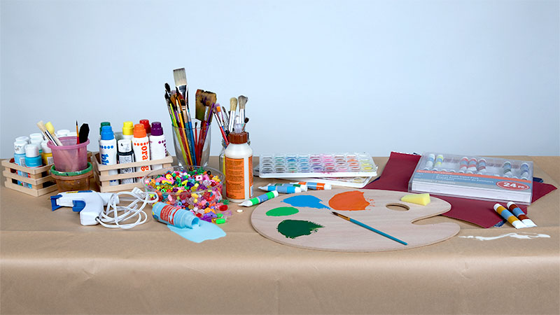 Paint, paintbrushes, glue, and other arts and crafts sit on table covered with paper. A DIY craft party is a great housewarming party theme idea.