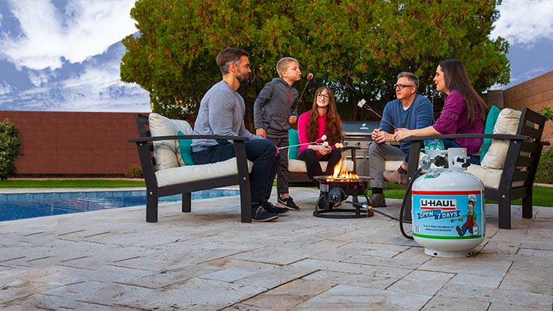 A family sits around a gas firepit roasting marshmallows in the backyard of their new home. A U-Haul propane tank lights the gas firepit.