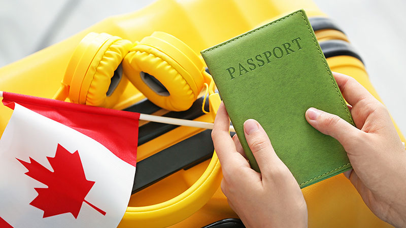 A moving abroad traveler carries their passport in their hands along with their yellow headphones, a yellow suitcase, and a Canadian flag.