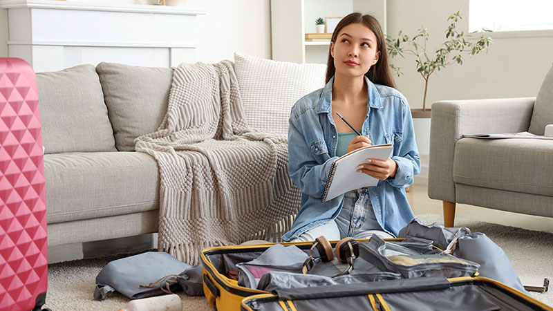 A woman thinks while writing a note down on her moving abroad checklist notebook. The woman sits on her knees in her living room while trying to pack her suitcases.