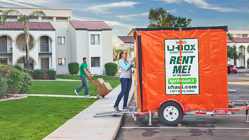 A woman places a basket into a U-Box storage container while a man in the background pushes a dolly with moving boxes toward the U-Box container.