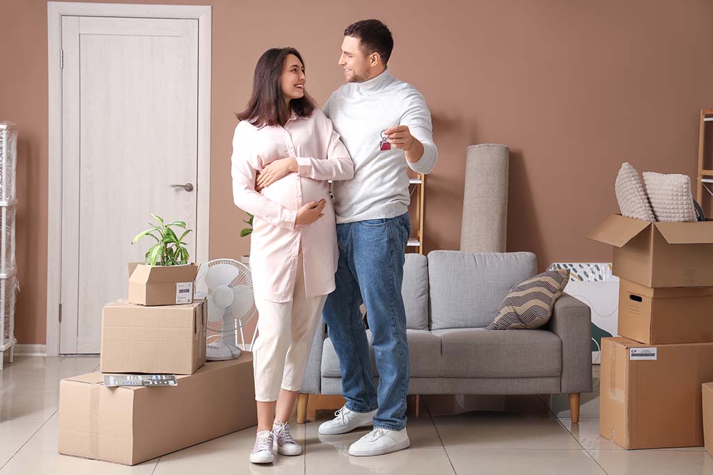 A couple stands together while the husband holds their keys to their new home. Moving boxes are on the ground surrounding the couple.