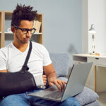 A man sits on a couch with a sling on his broken right arm while looking at his computer.