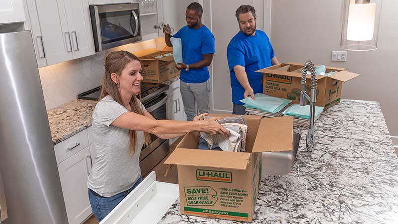 Two Moving Helpers pack plates, glasses, and stemware into glass packing kit with a moving box while the customer packs dish clothes into a box in the customer’s kitchen.