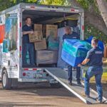 Two Moving Helpers work together down the ramp of a U-Haul truck rental as they carry the customer’s couch. Another labor-only mover stands in the U-Haul truck rental waiting for his turn to go down the ramp.